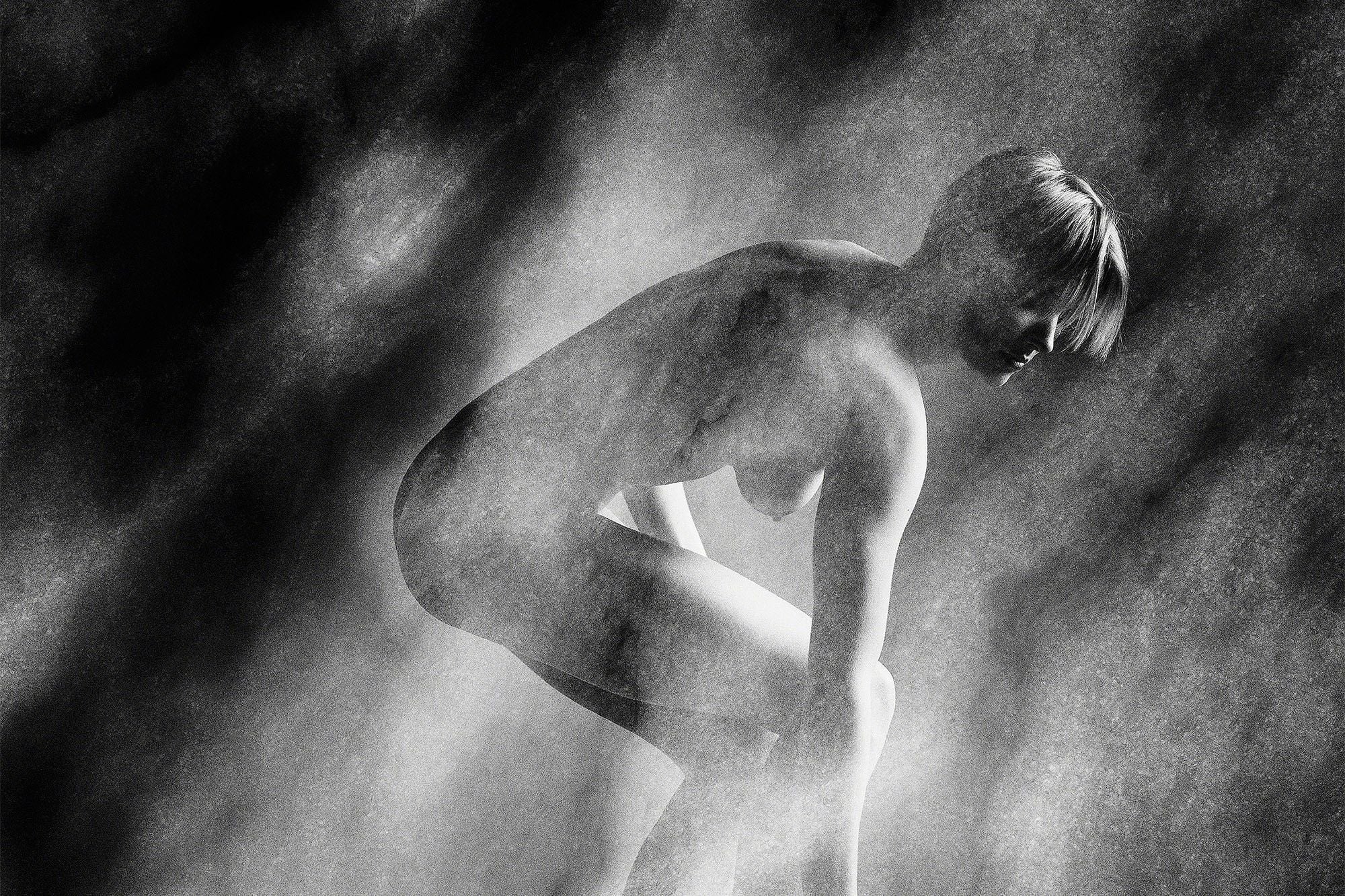 Black and white study of a nude figure crouching forward, streaked with soft light against a dark textured background